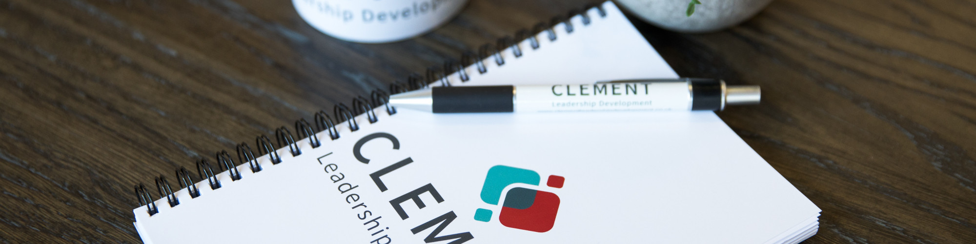 A notepad and pen with the word Clement on, with a plant and coffee mug nearby